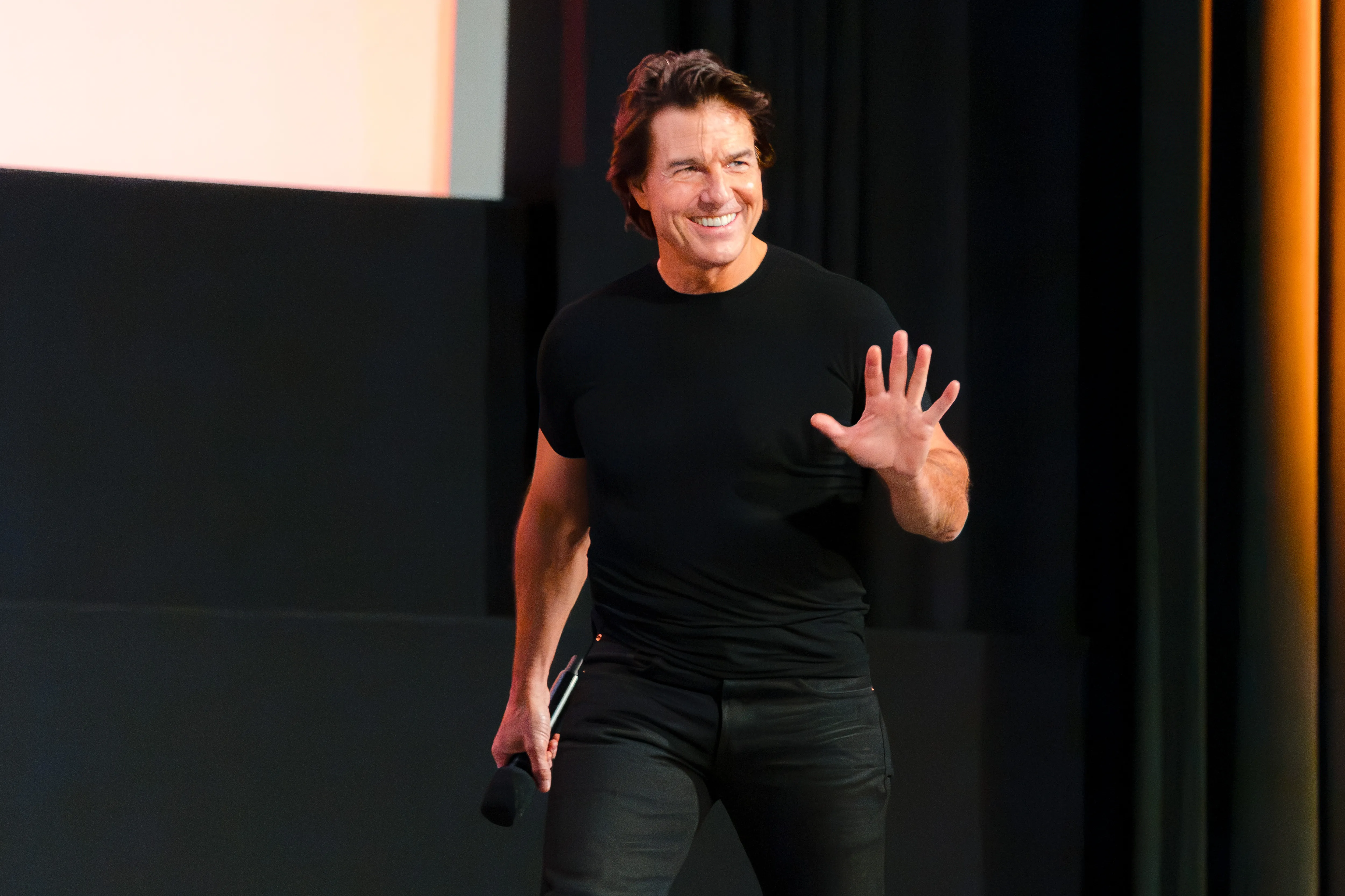 Tom Cruise smiling and waving his left hand while holding a microphone at the Digger Trailer Launch Event.