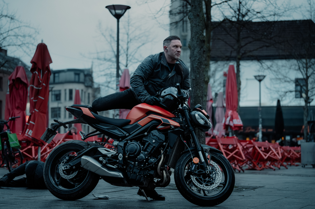 Tom Hardy as Harry Da Souza perched on an orange motorcycle with a person lying on the ground next to the back wheel.