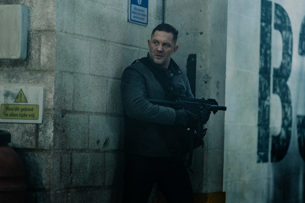 Tom Hardy as Harry Da Souza holding a gun and looking over his shoulder.