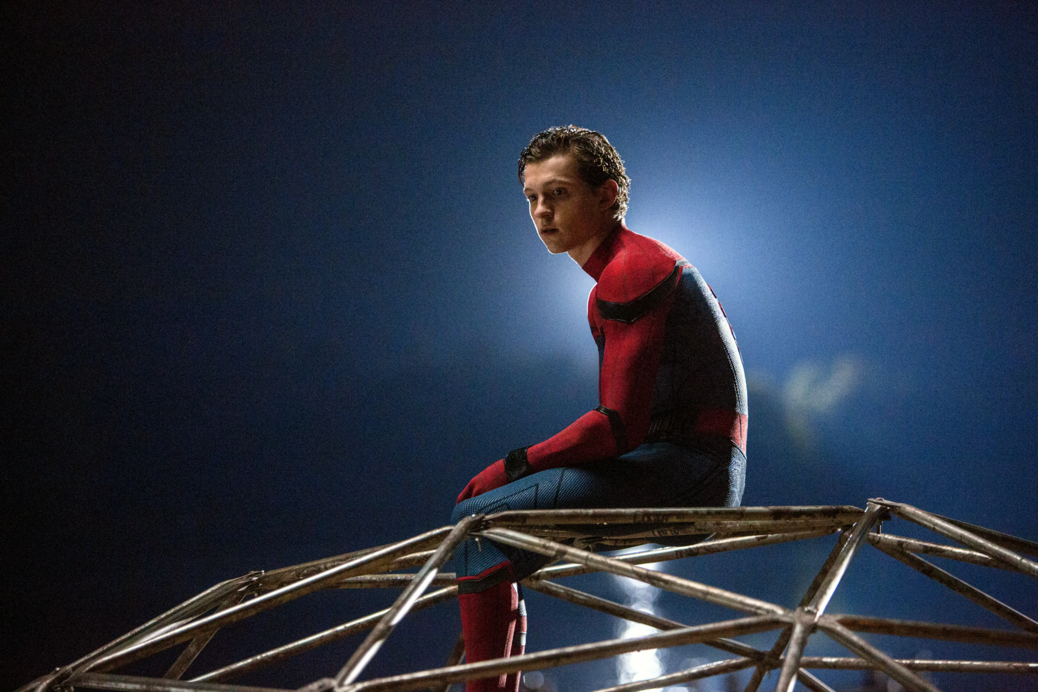 Tom Holland as Spider-Man sitting on a metal structure in a spotlight.