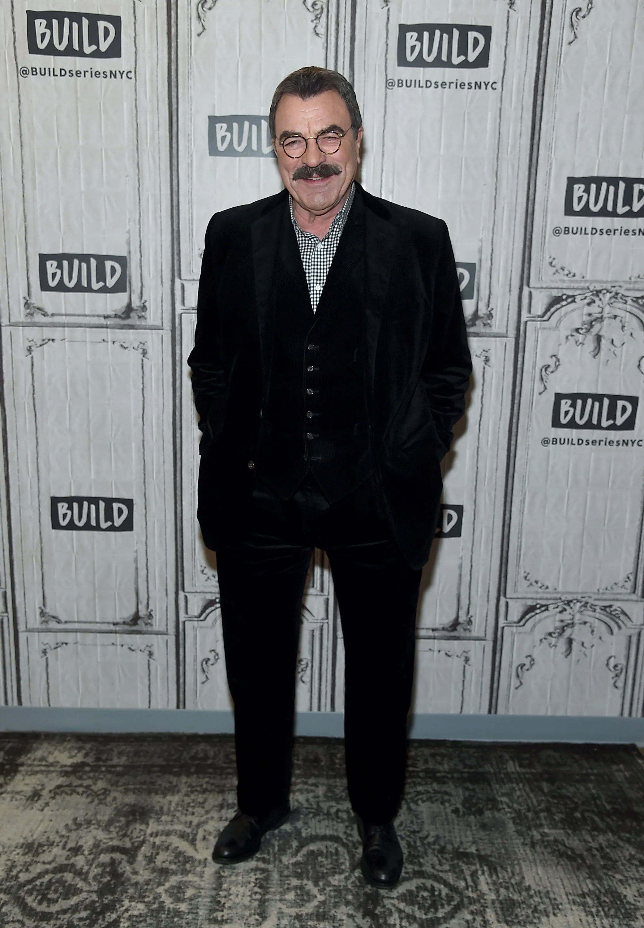 Tom Selleck smiling in a black velvet suit, a white and black checkered shirt, and round glasses, standing against a backdrop of white panels with the word "BUILD" and "@BUILDseriesNYC" repeated.
