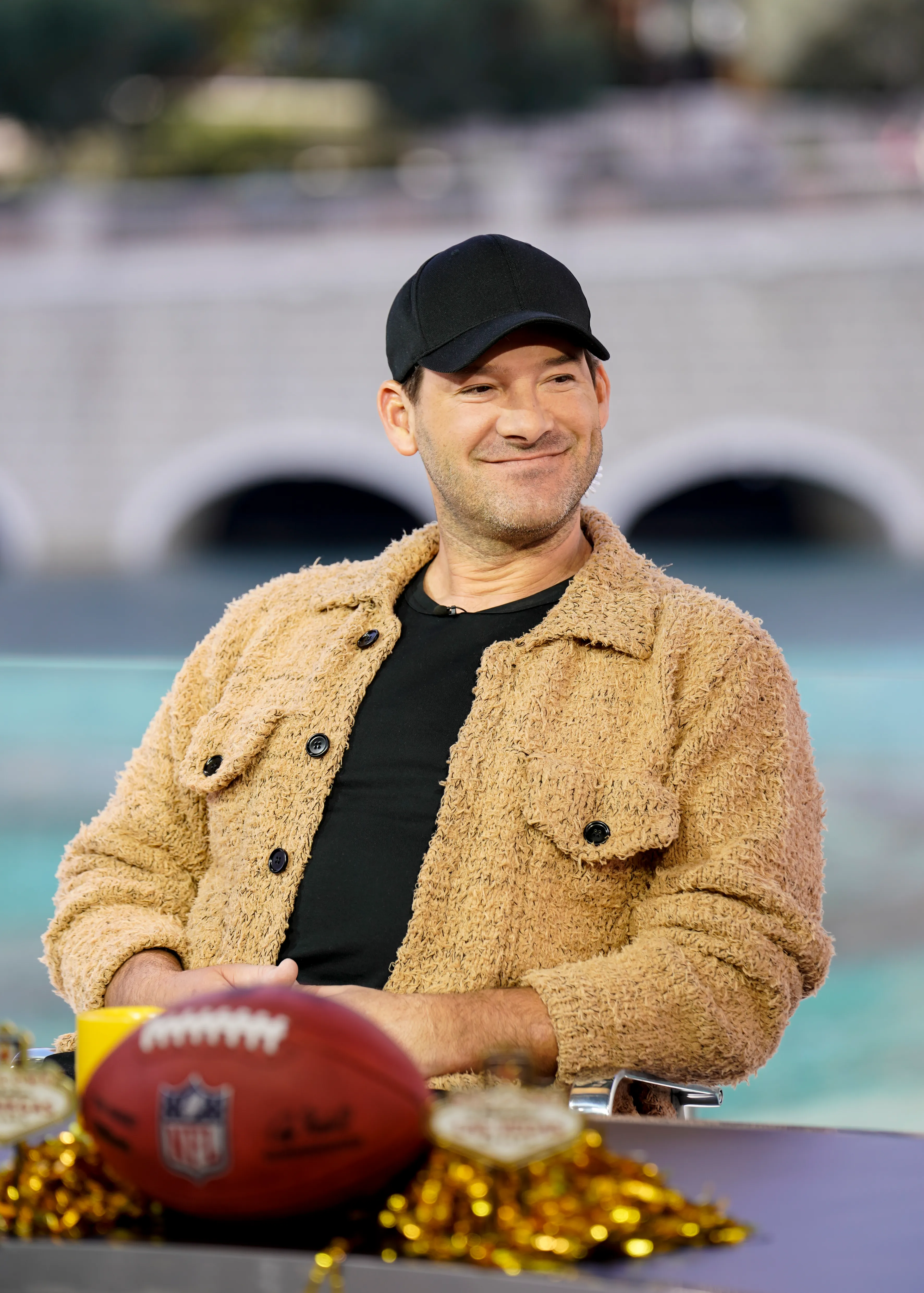 Tony Romo, wearing a black baseball cap and a textured tan jacket, smiles while sitting at a table with an NFL football.