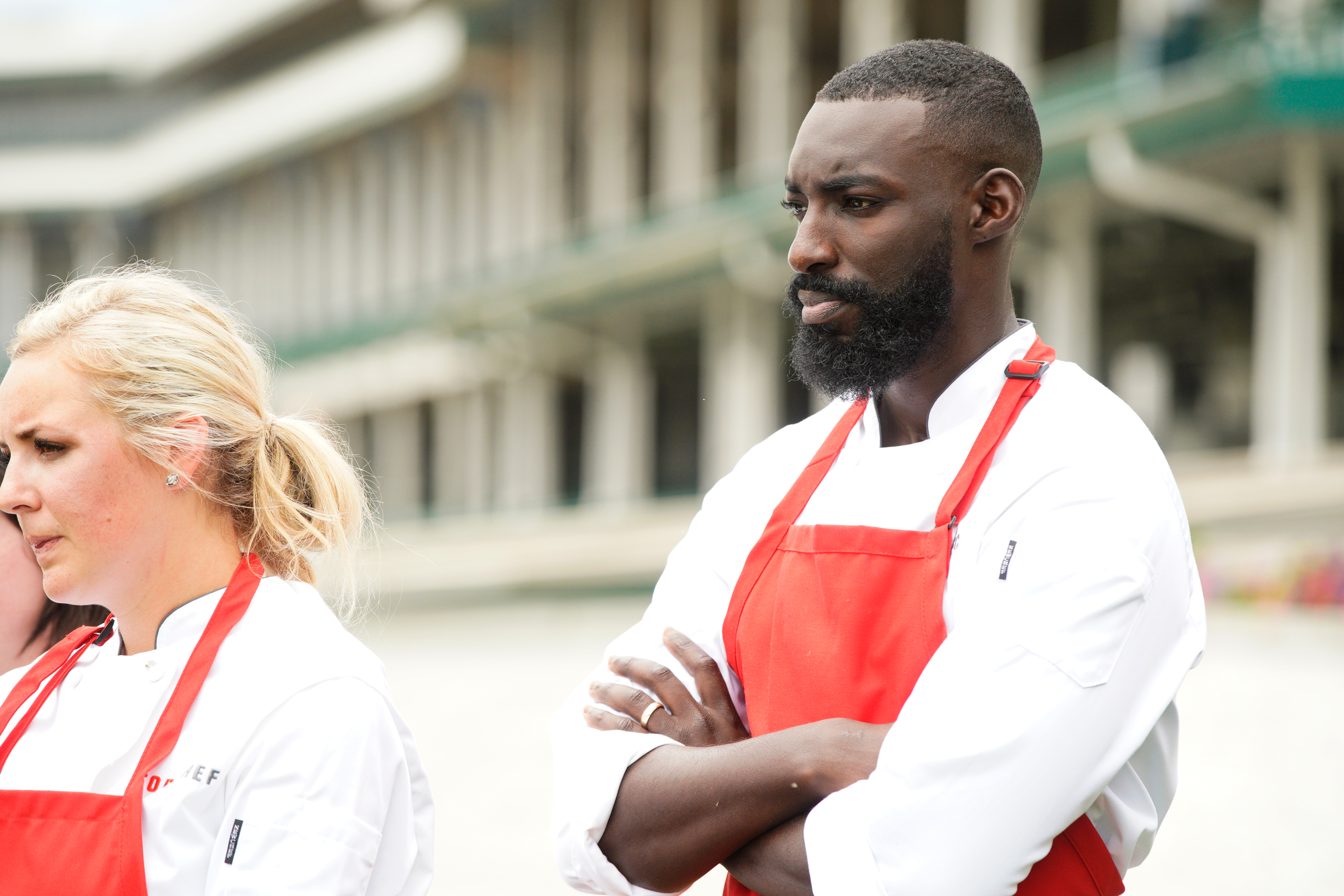Chef Eric Adjepong and Kelsey Barnard in white chef's coats and red aprons stand outdoors.