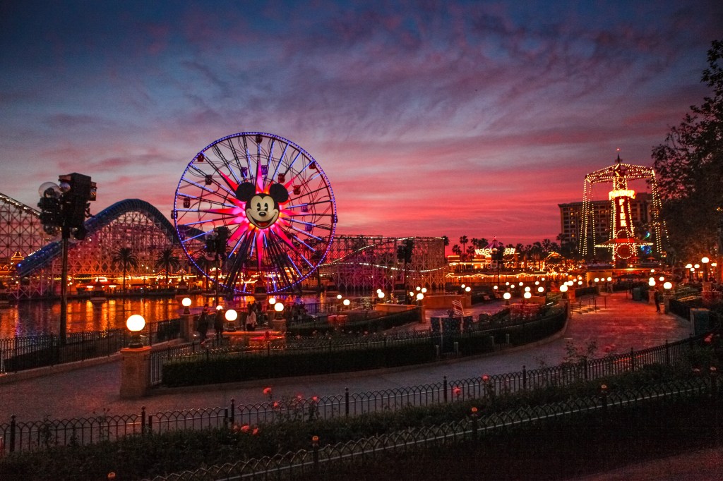 Paradise Pier at Disney's California Adventure at sunset, featuring the illuminated Ferris wheel with Mickey Mouse in the center, and a roller coaster in the background.