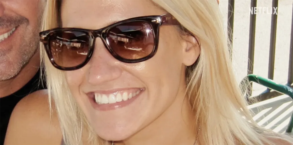 A woman with blonde hair and sunglasses smiles, with a man's face partially visible beside her.
