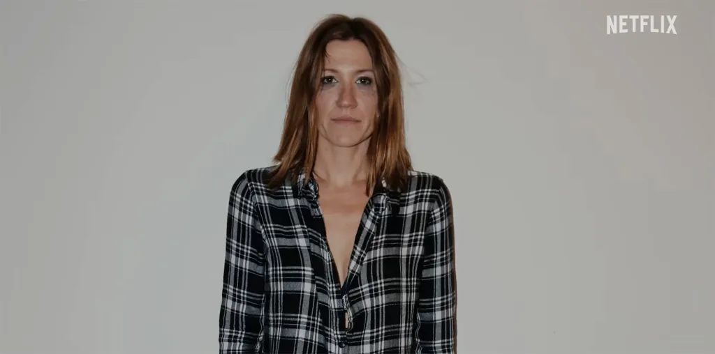 A woman with disheveled hair and dark makeup under her eyes wears a black and white plaid shirt.