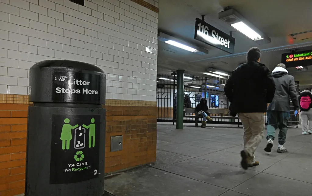 A black trash can with "Litter Stops Here" and recycling instructions stands against a tiled wall in the 116th Street subway station, with people walking in the background.
