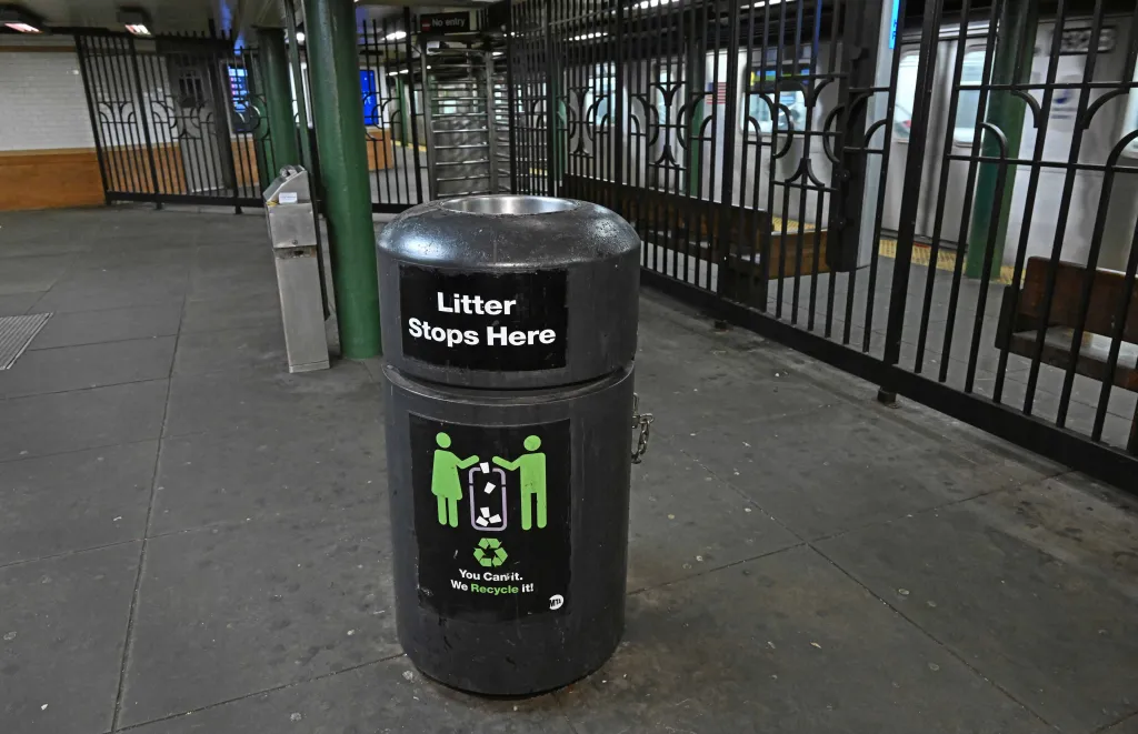 A black garbage can with the text "Litter Stops Here" and "You Can It. We Recycle It!" in a subway station with a train on the tracks behind a fence.