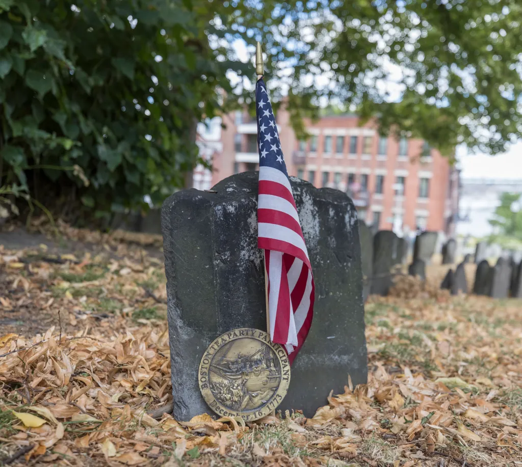 A small American flag and a medal commemorating the Boston Tea Party are placed against a tombstone in Copp's Hill Burying Ground.