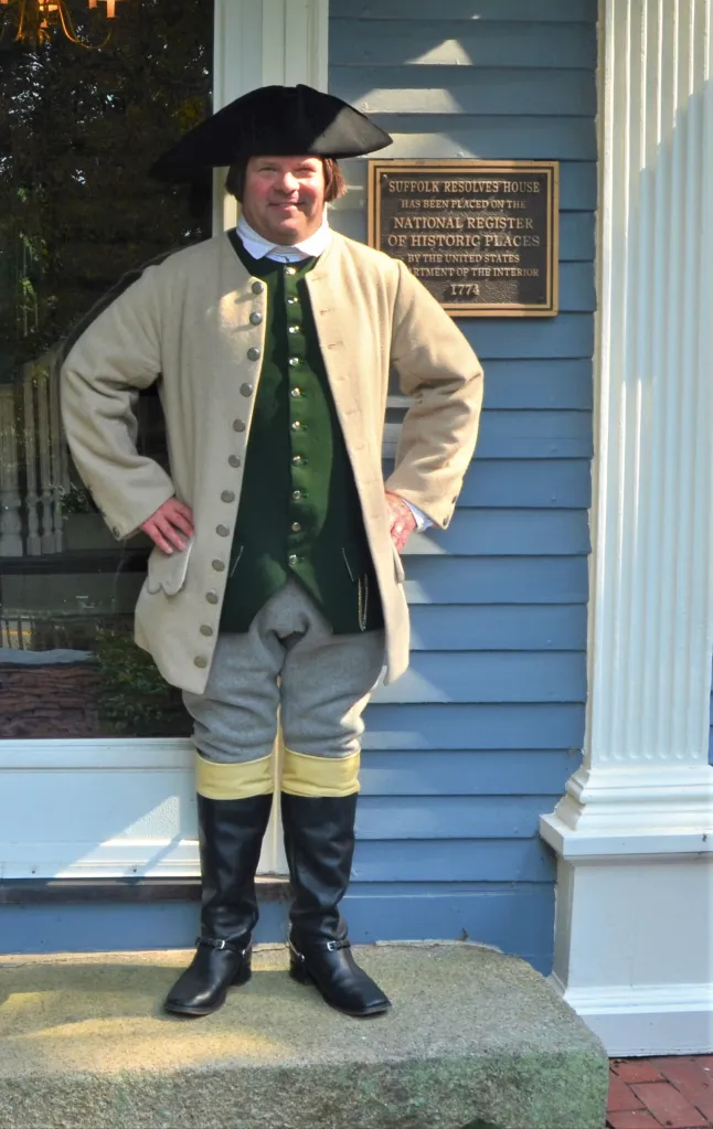 Paul Revere reenactor Michael Lepage in colonial attire standing next to a house with a plaque that reads "Suffolk Resolves House, 1774."