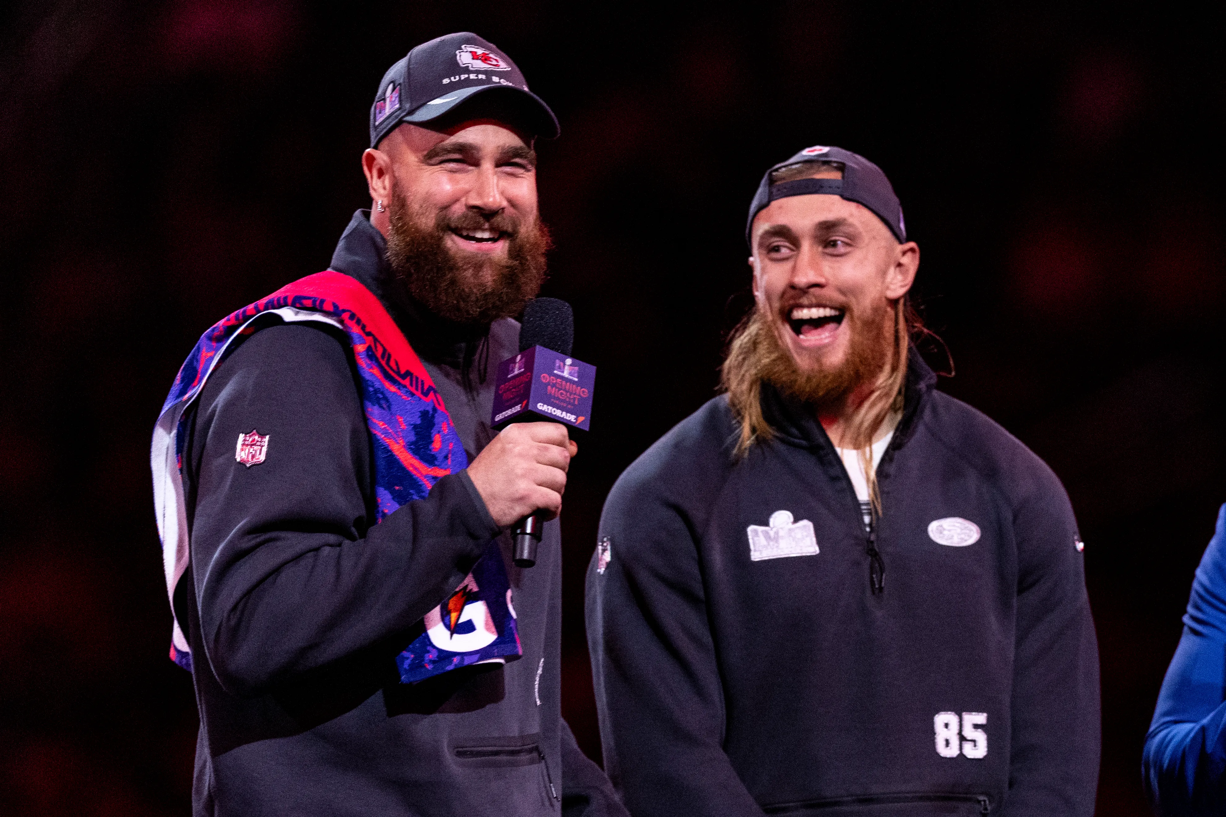Travis Kelce holding a microphone and smiling next to George Kittle.