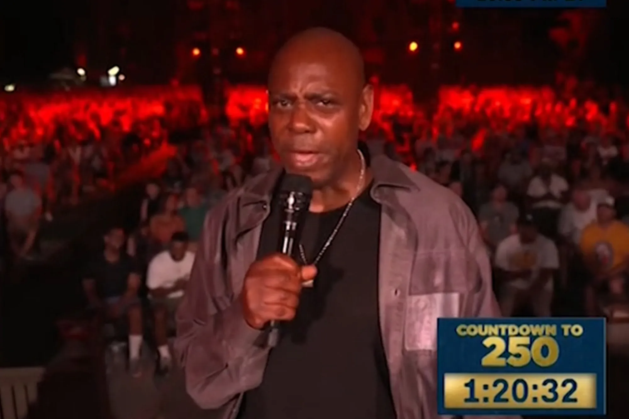 Dave Chappelle speaks into a microphone on stage in front of a red-lit crowd.