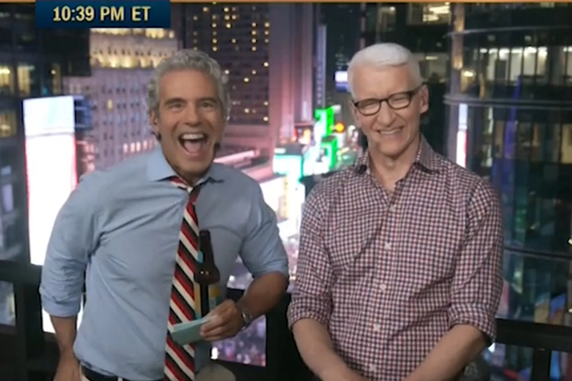 Andy Cohen and Anderson Cooper laughing with a city background.