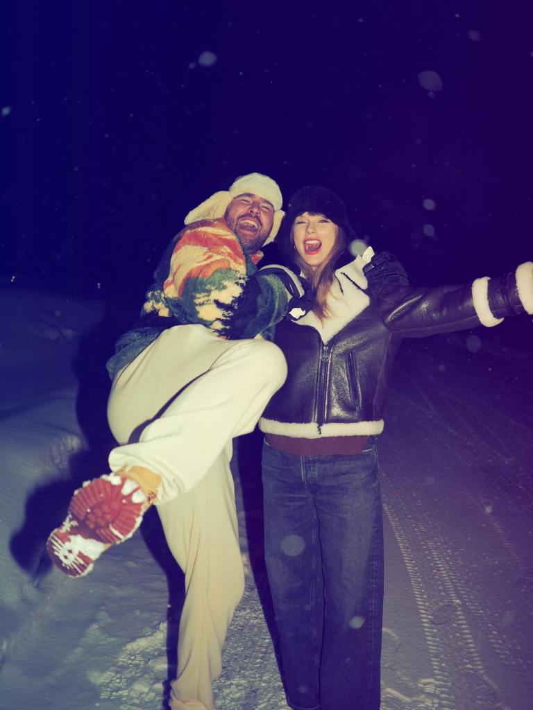 Taylor Swift holds Travis Kelce as he lifts a leg, both smiling and shouting in the snow at night.