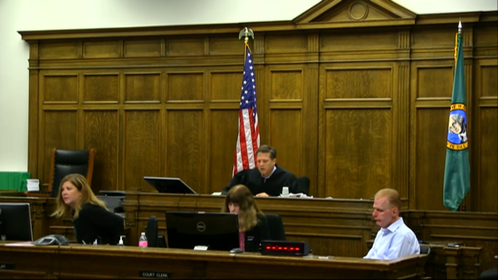 A courtroom with a judge, a clerk, and a man in a light blue shirt.