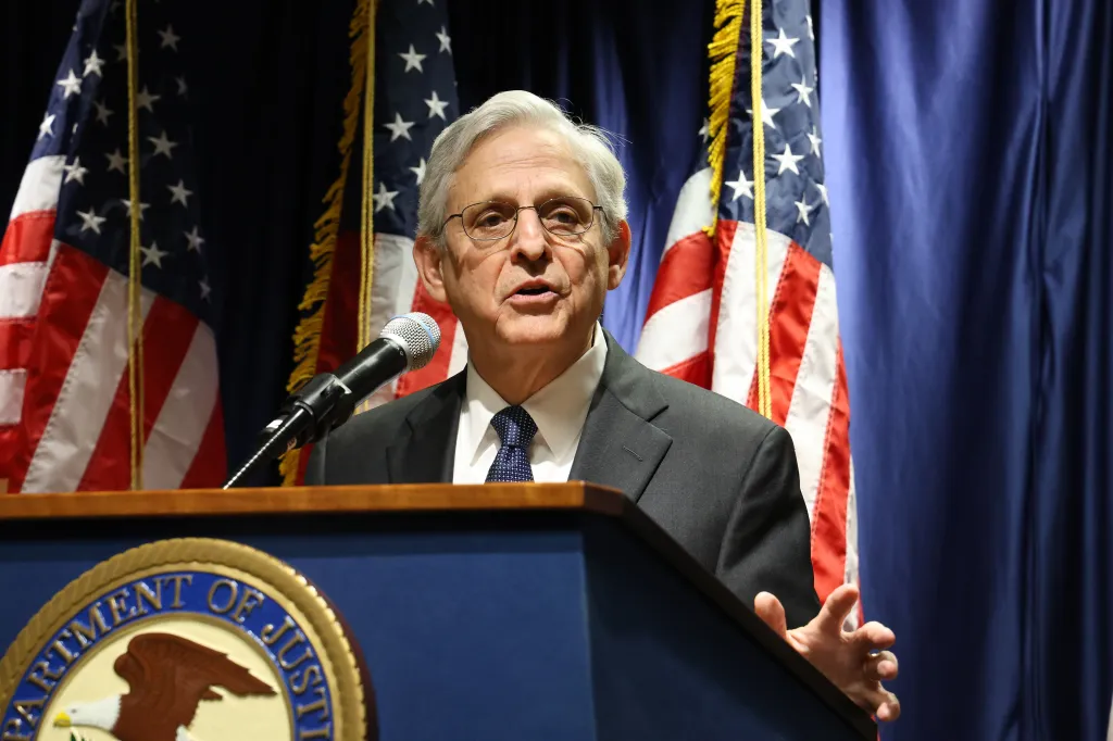 U.S. Attorney General Merrick Garland speaking at a podium with two American flags in the background.