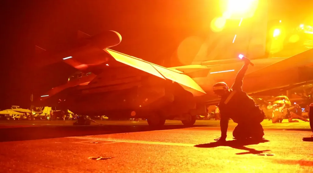 A US Navy crew member kneeling on a brightly lit flight deck, guiding an F/A-18 Super Hornet fighter jet at night.