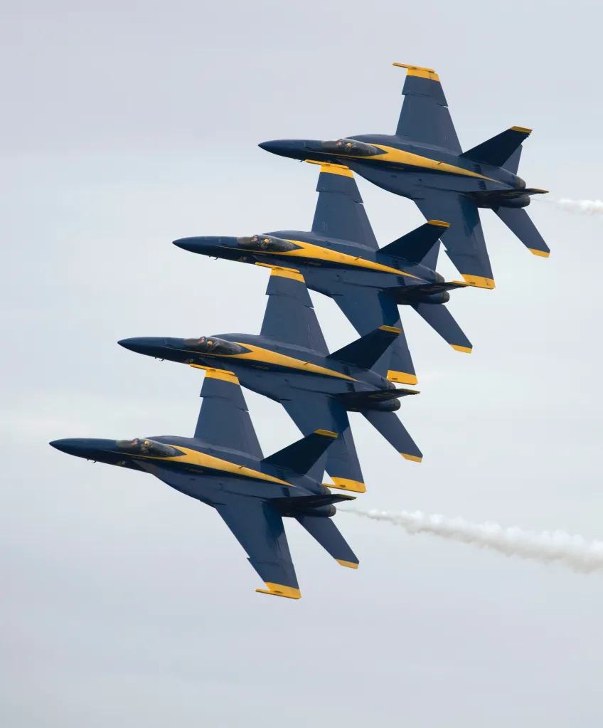 Four US Navy Blue Angels jets flying in formation, leaving white smoke trails.