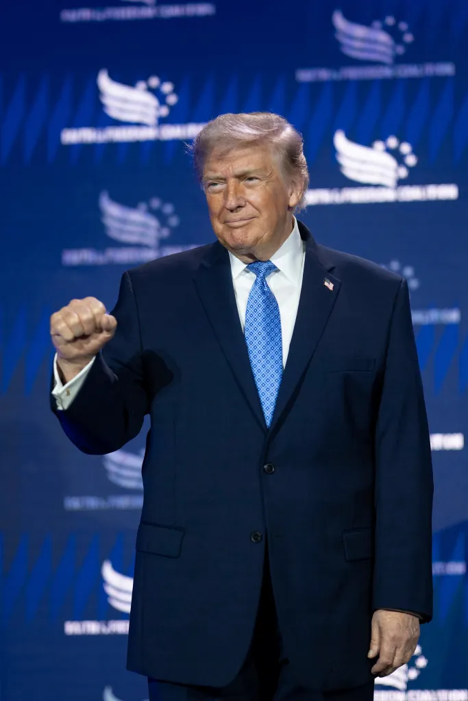 Donald Trump, wearing a dark blue suit and light blue tie, makes a fist with his right hand at a podium in front of a blue background with white logos.