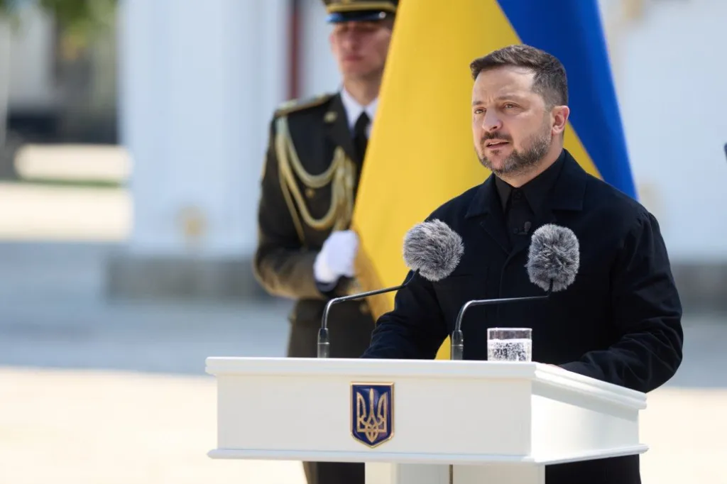 Ukrainian President Volodymyr Zelenskyy speaking at a podium with microphones, a glass of water, and the Ukrainian coat of arms, with a Ukrainian flag and a soldier in the background.