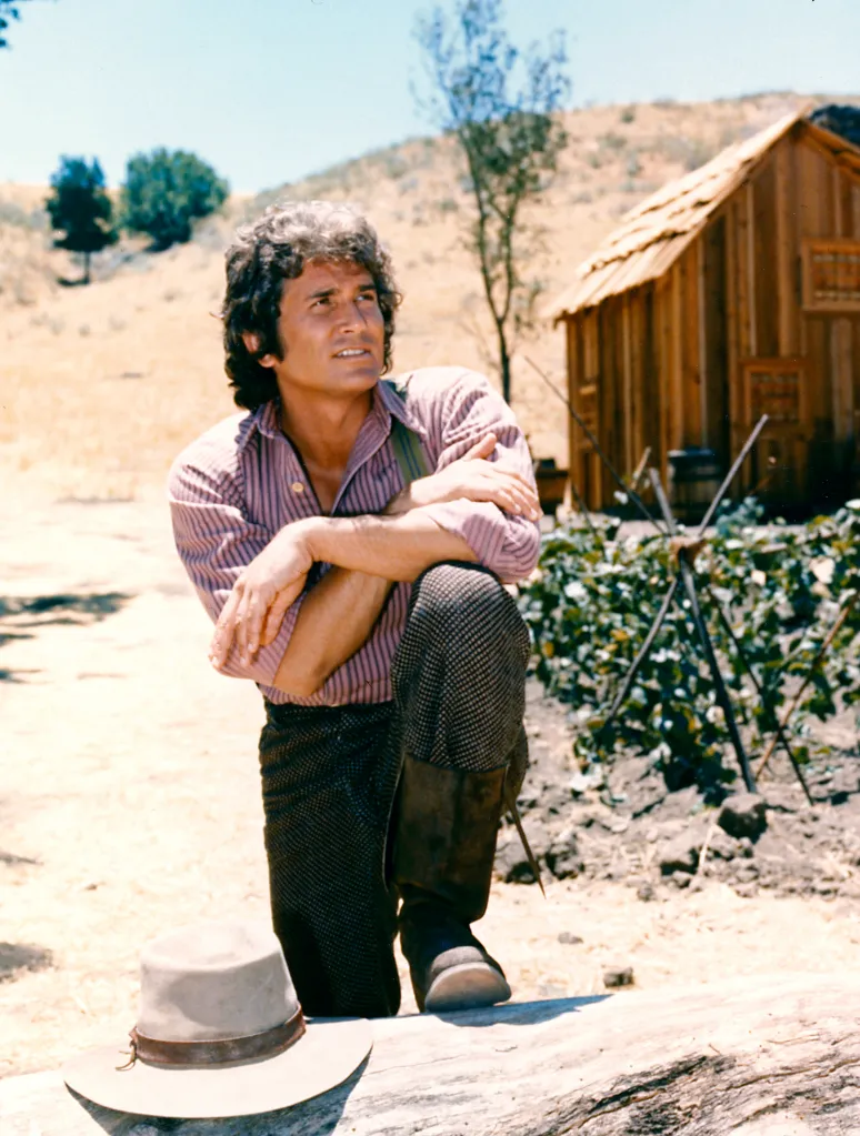 US actor Michael Landon as Charles Ingalls from 'Little House on the Prairie' posing in a striped shirt and patterned pants, with a log cabin in the background.