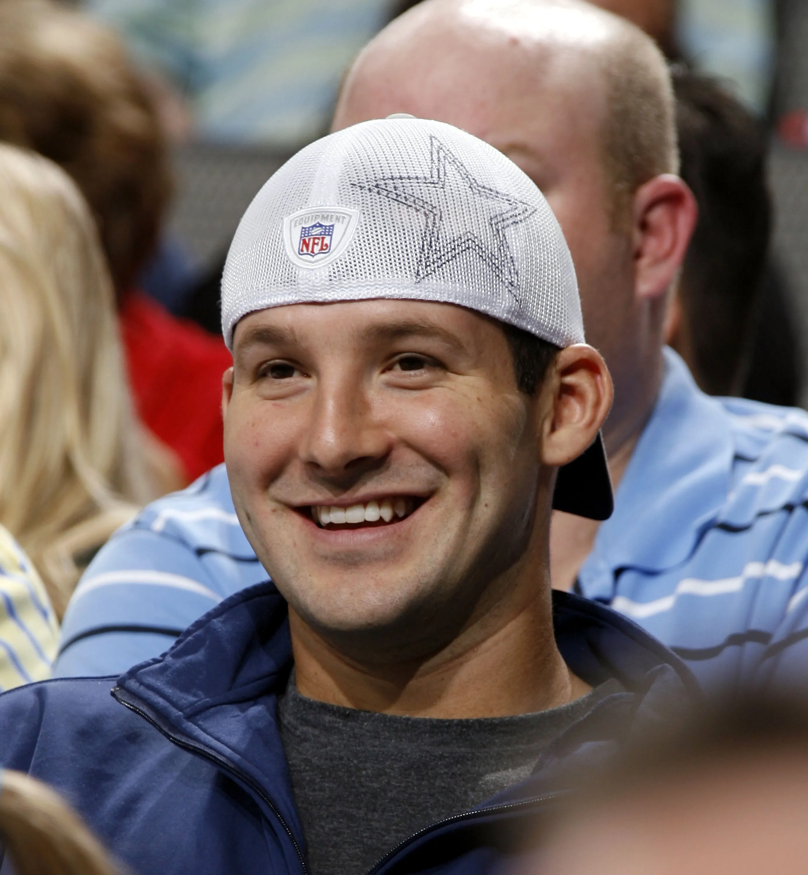 Tony Romo smiling, wearing a white Dallas Cowboys cap and a blue jacket.