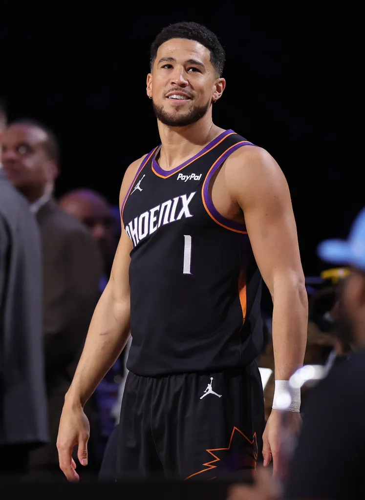Devin Booker smiling while wearing a black Phoenix Suns jersey with "PHOENIX" and the number "1" on the front.