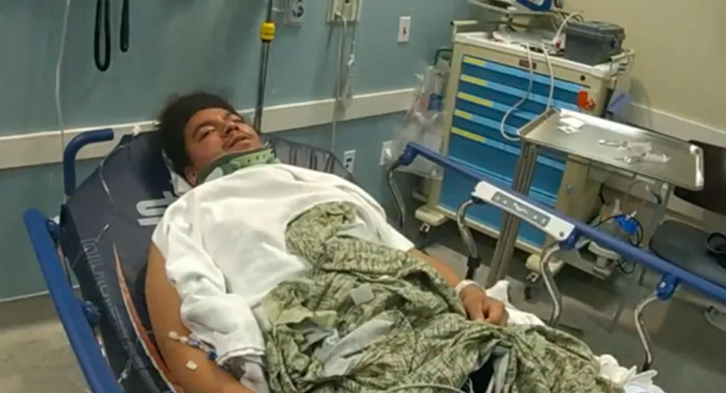 A man lying in a hospital bed with a neck brace and an IV in his arm, with bruising on his face, chest, and arms, and swelling on his forehead.