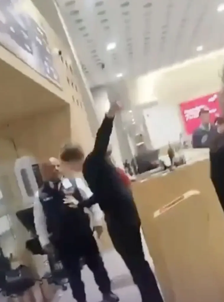 A blurred still image from a video shows a man in a dark top with his arm raised, possibly in a struggle, with other people and airport structures visible in the background.