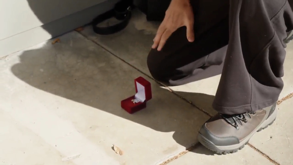 A person on one knee with a red ring box open on the ground in front of them.