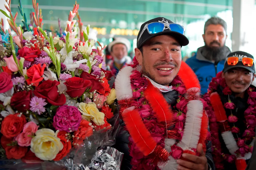 Nirmal Purja, with a scarred nose, smiling while wearing a hat, sunglasses, and a floral garland around his neck, stands next to a large bouquet of flowers with other men behind him.