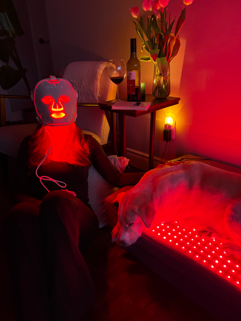 A person with a red light therapy mask on sits next to a dog resting on a red light therapy mat.