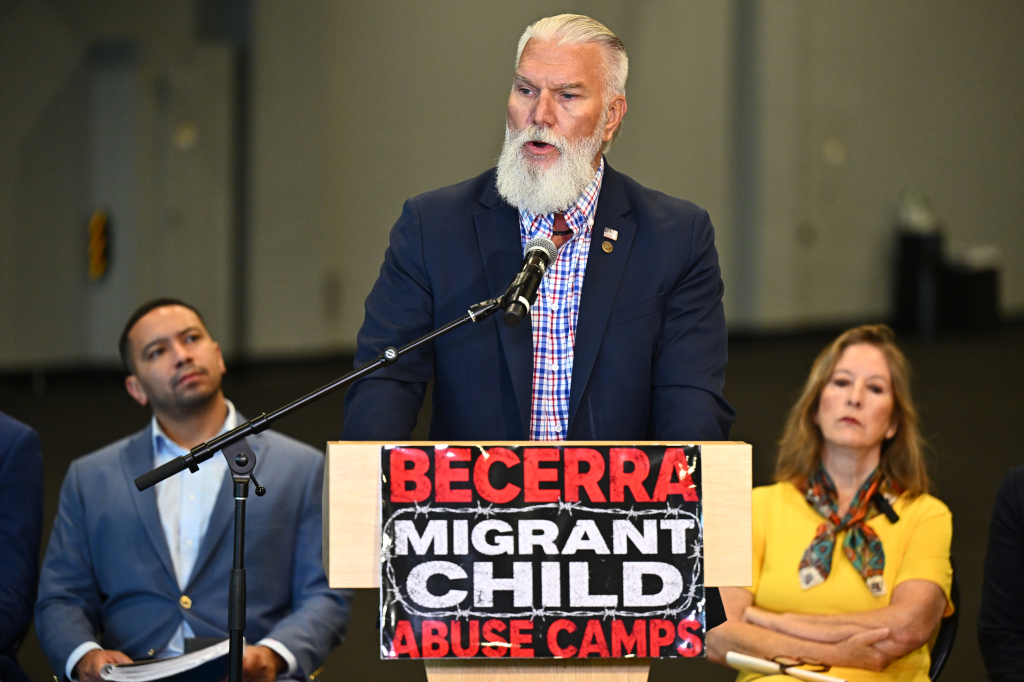U.S. Border Patrol Chief Chris Clem speaks at a news conference from a podium adorned with a sign reading, "BECERRA MIGRANT CHILD ABUSE CAMPS."
