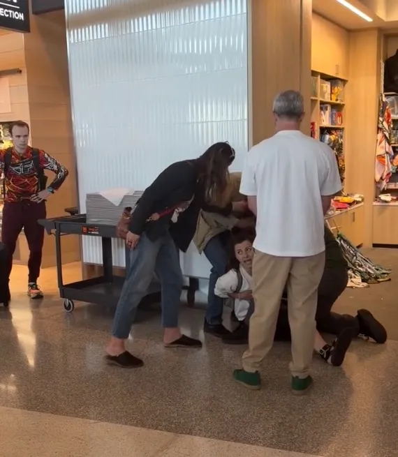 Multiple people gathered around a person on the floor at an airport, with another person watching from a distance.