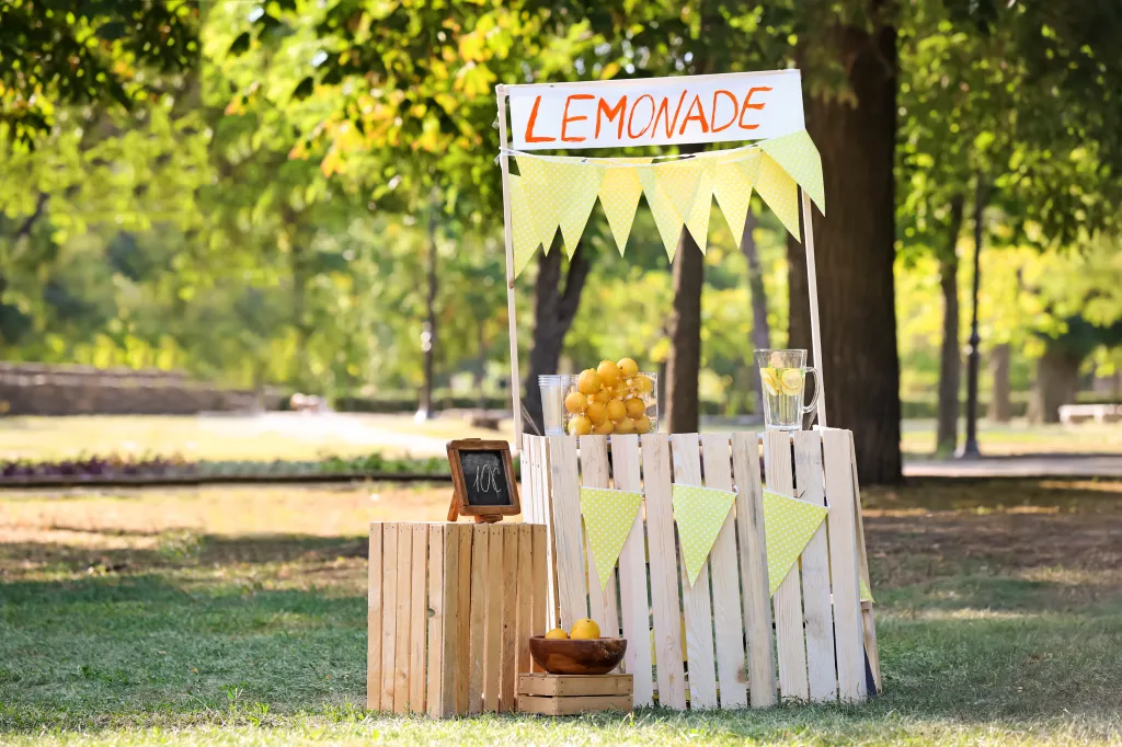 A wooden lemonade stand with a "LEMONADE" sign, decorated with yellow polka-dot bunting, a chalkboard sign, lemons, and a pitcher of lemonade, set up outdoors on a sunny day.