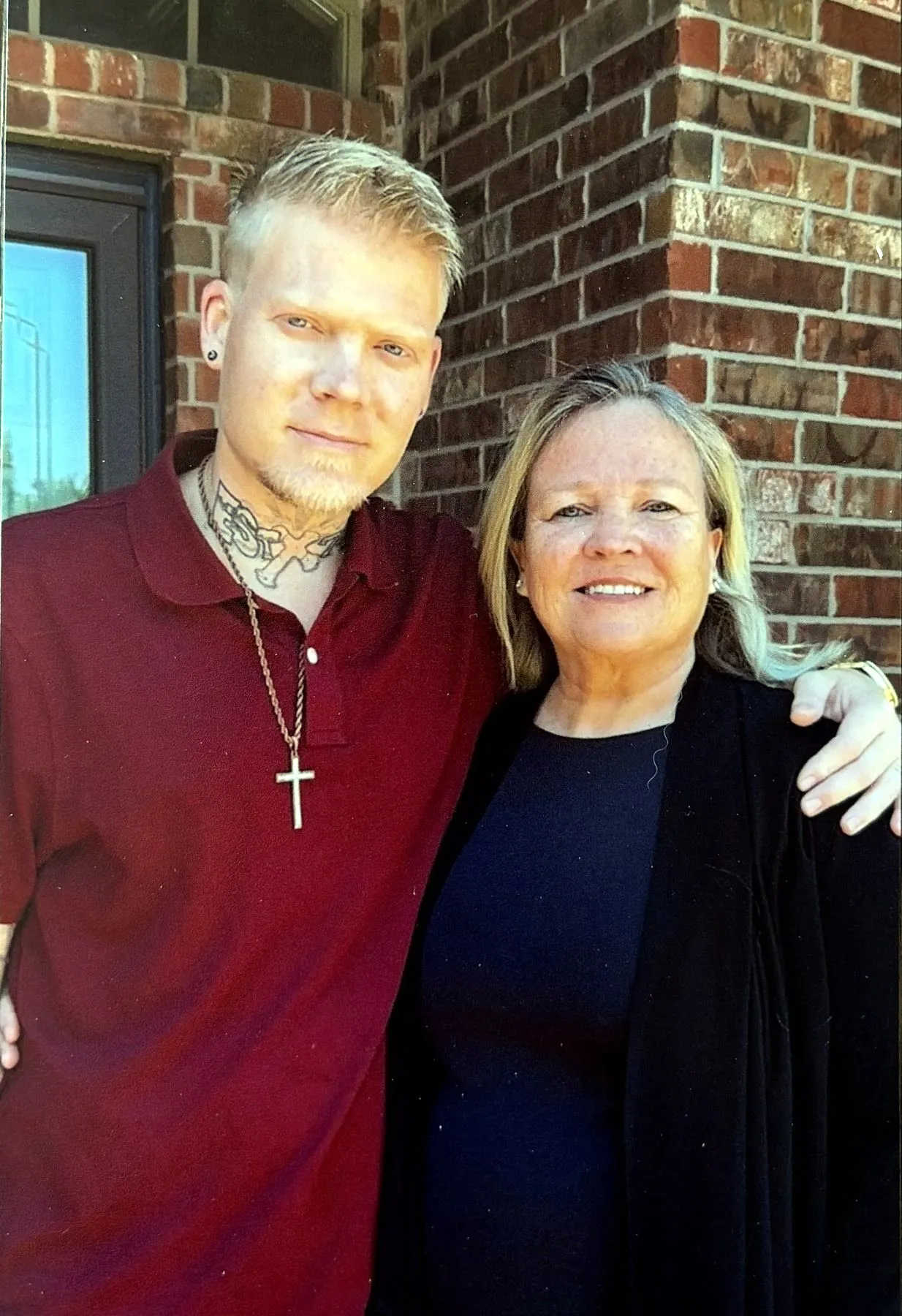 A light-skinned man with blonde hair and a beard, wearing a maroon shirt with a cross necklace, and a light-skinned woman with blonde hair, wearing a black top and jacket, stand smiling with their arms around each other.