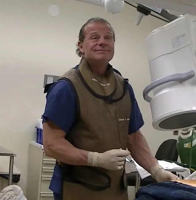 A doctor named Gabriele Jasper, MD, wearing a lead apron and surgical gloves, stands next to medical equipment.