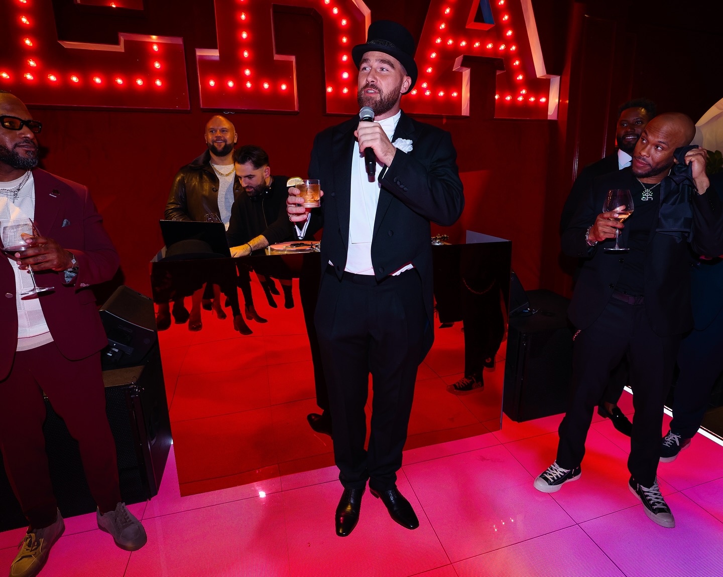 Travis Kelce speaking into a microphone while wearing a top hat and tuxedo at a party.