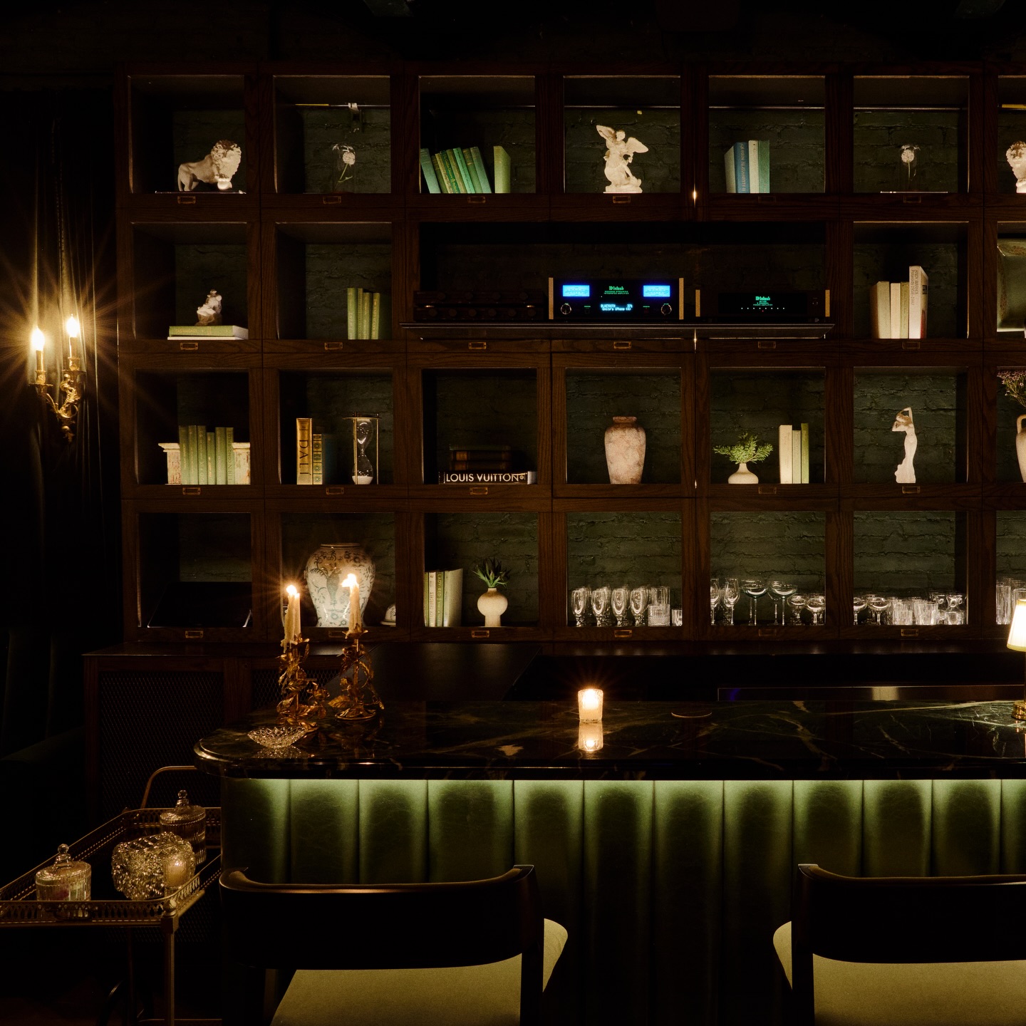 A dimly lit bar at Dandelion in West Village, NYC, with a green upholstered bar front and a large display cabinet filled with decorative items and books.