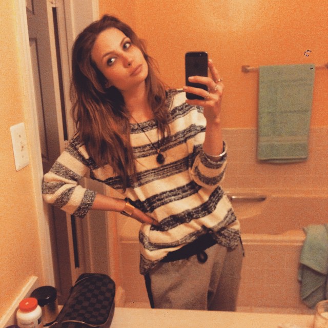 Daveigh Chase takes a mirror selfie while wearing a striped shirt and grey sweatpants.