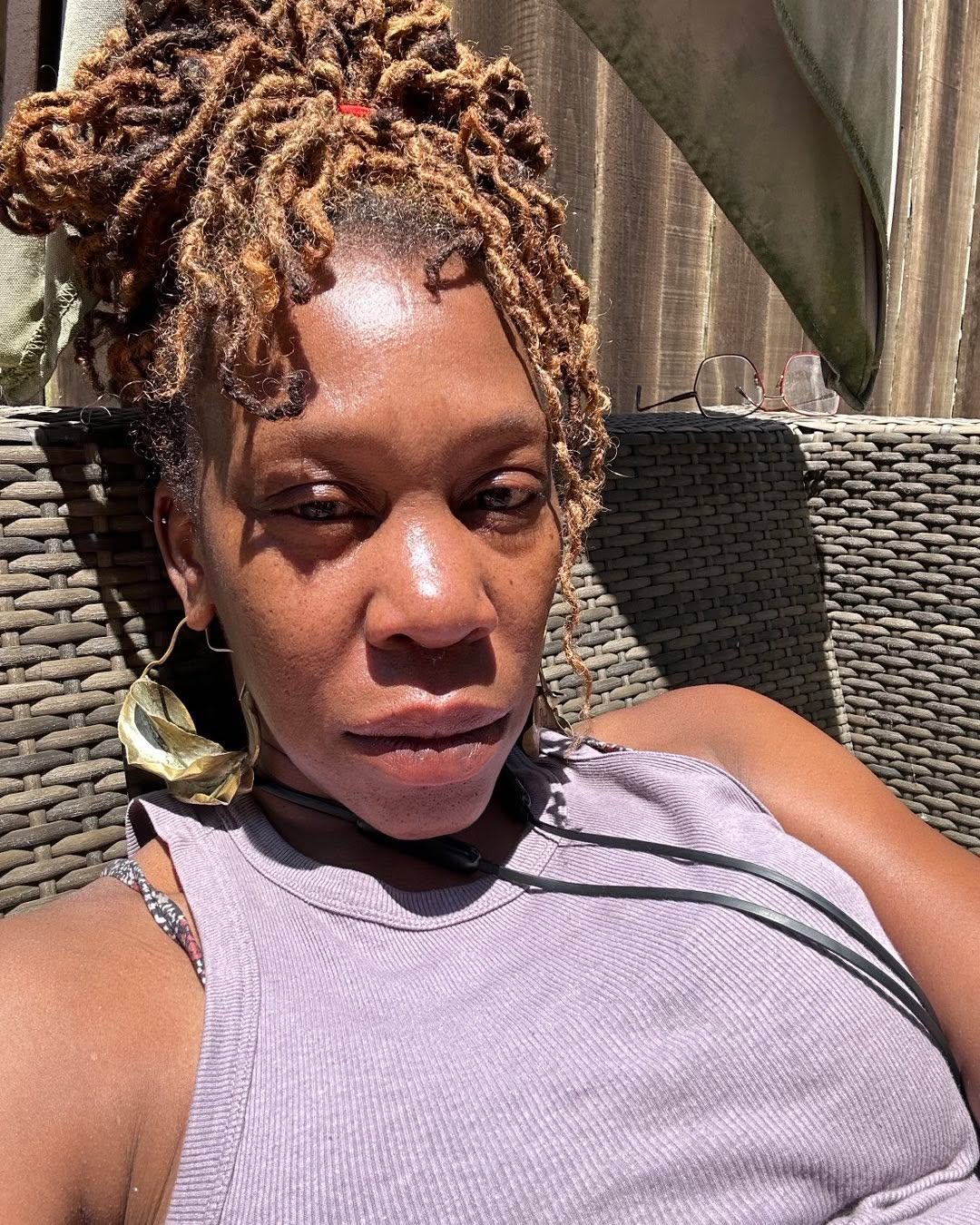 A woman with light brown dreadlocks and large gold earrings wearing a lilac tank top.