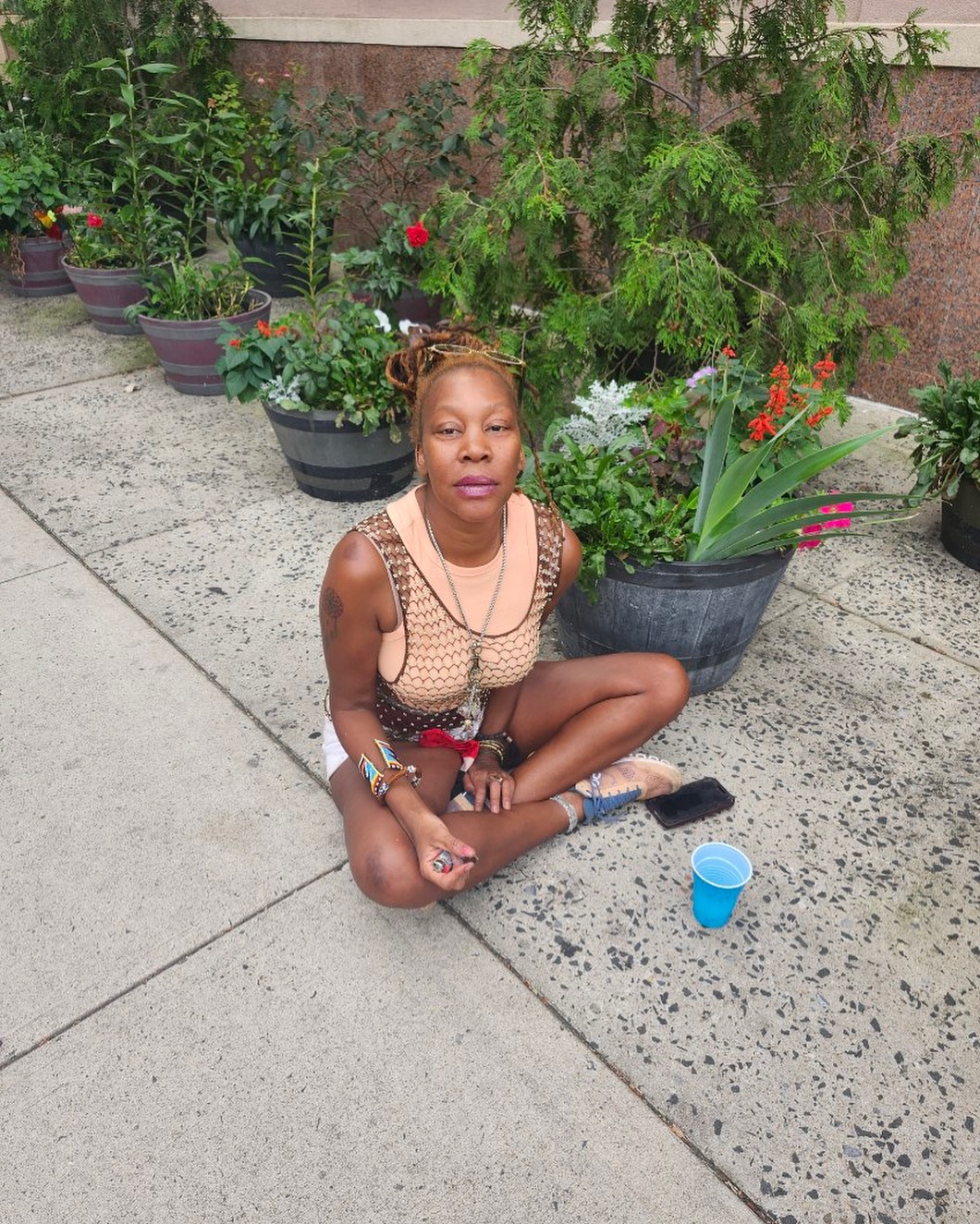 A woman with dreadlocks and sunglasses on her head, wearing a mesh top and white shorts, sits cross-legged on a sidewalk among potted plants.