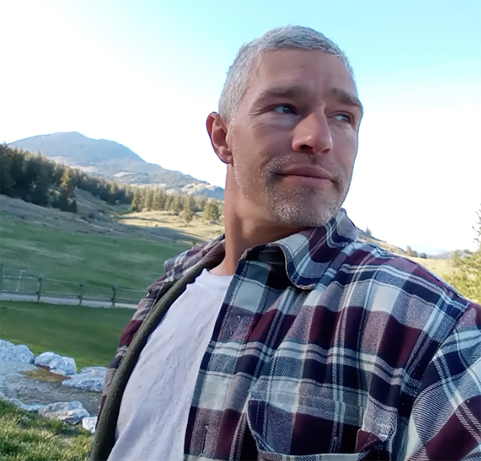 Man with graying hair and beard looks to his right, wearing a plaid shirt and white T-shirt with mountains and a green field in the background.