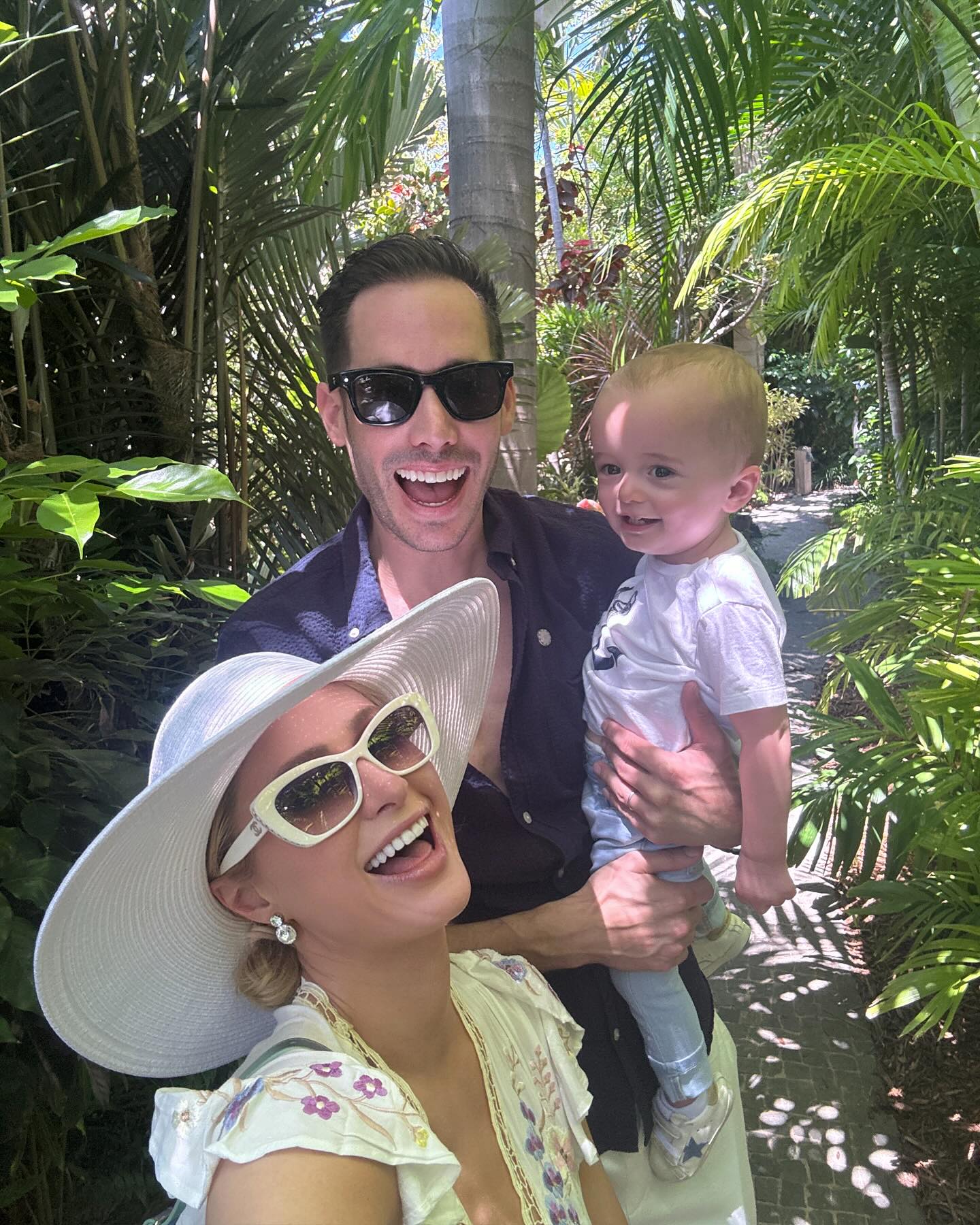 Paris Hilton, Carter Reum, and their son Phoenix smiling outdoors in a tropical setting.
