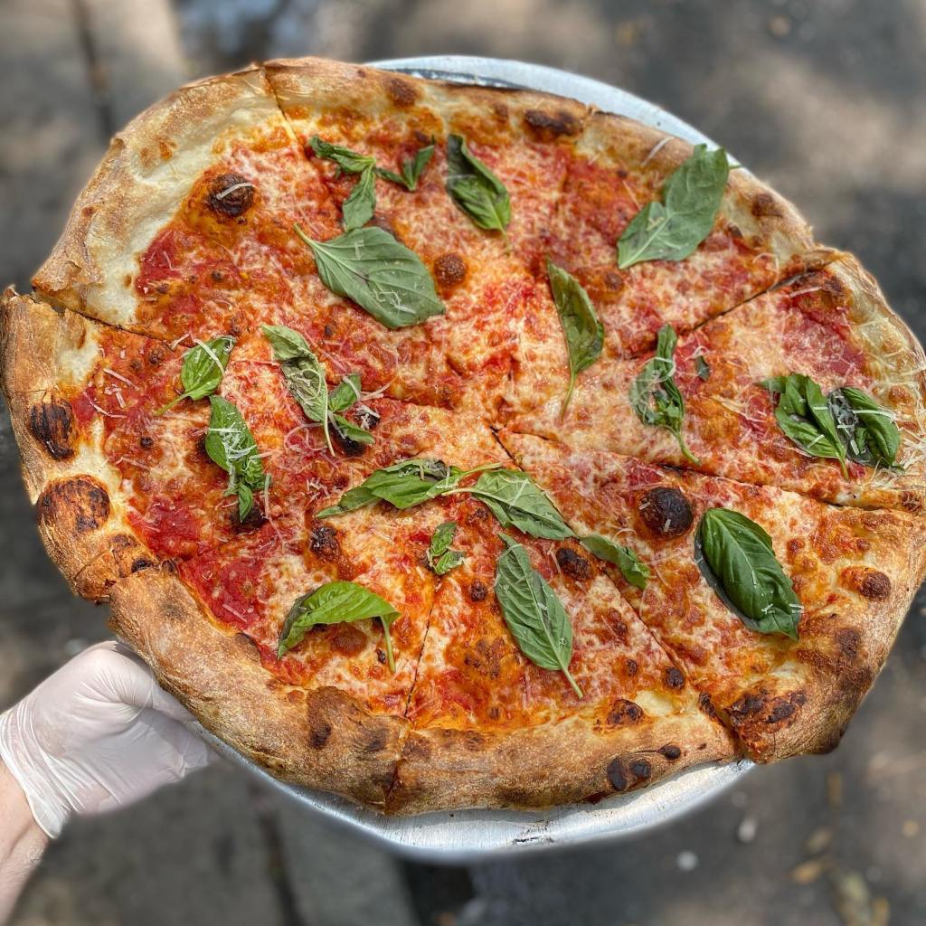 A pizza from Mama's Too, with red sauce, cheese, and basil leaves, being held by a gloved hand.