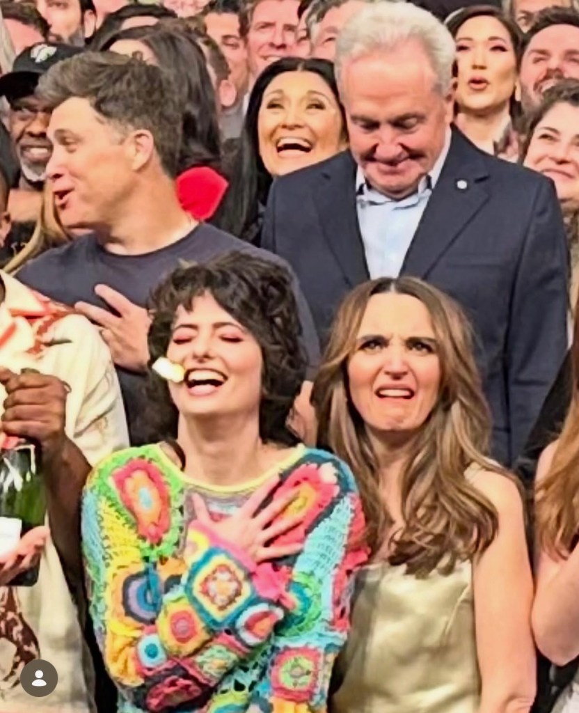 Chloe Fineman in a colorful crocheted top, holding a marshmallow in her mouth, laughs with fellow cast members.