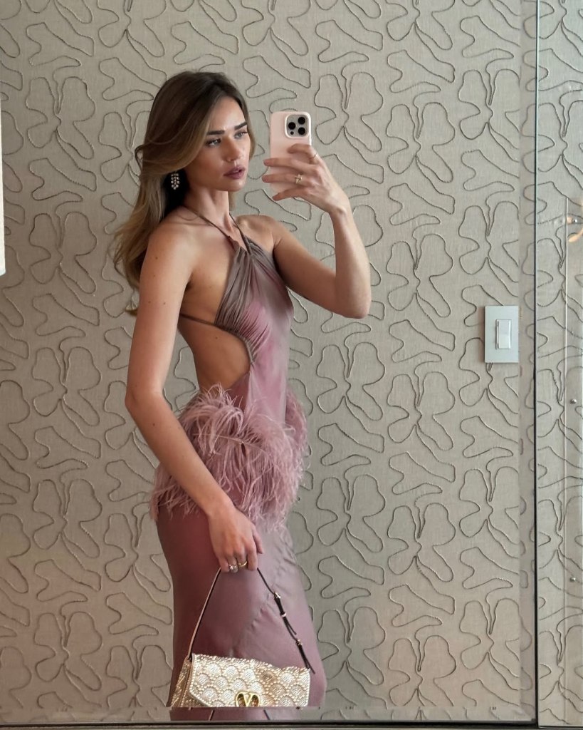 A woman in a mauve halter-neck dress with feather details on the side poses for a mirror selfie, holding a golden purse.