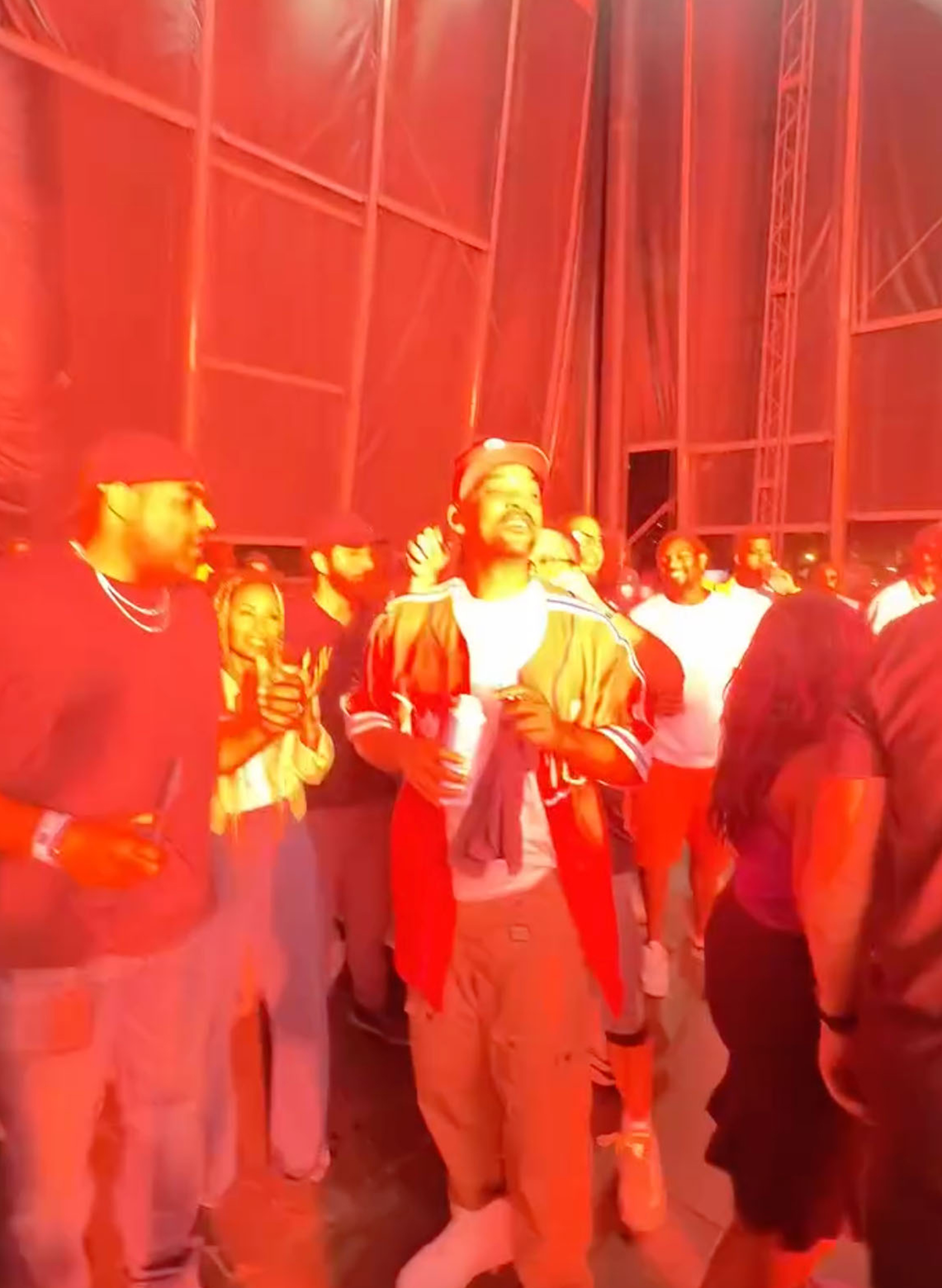 Will Smith in a baseball jersey with a white shirt and khaki pants, holding a cup in his left hand, surrounded by a crowd under red lighting.