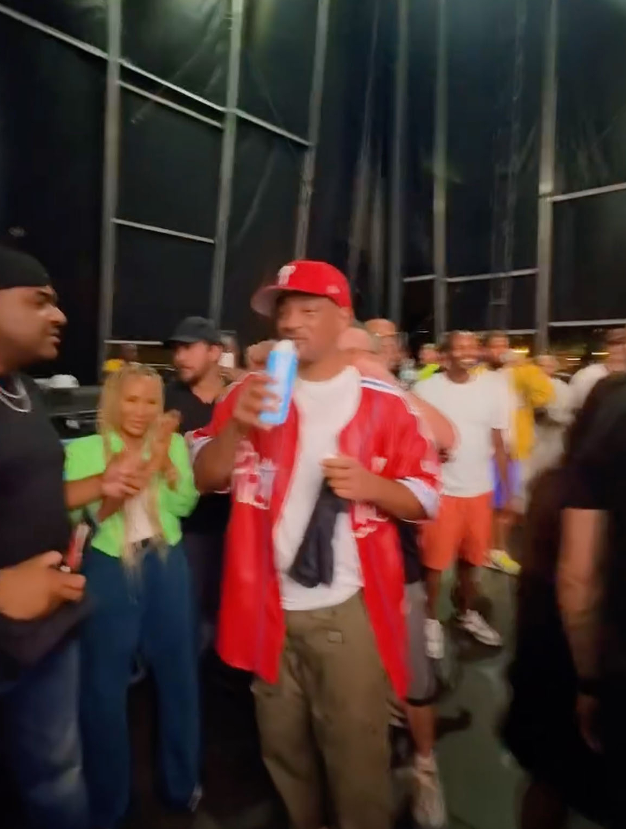 Will Smith holding a light blue bottle and wearing a red hat, a white t-shirt, and a red jacket, surrounded by people.