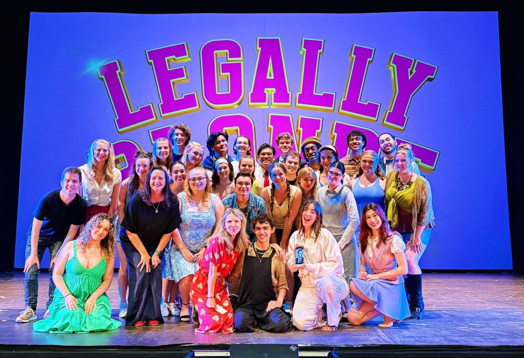 A large group of smiling people stand and kneel on a stage in front of a blue backdrop with the pink and yellow outlined words "LEGALLY BLONDE".